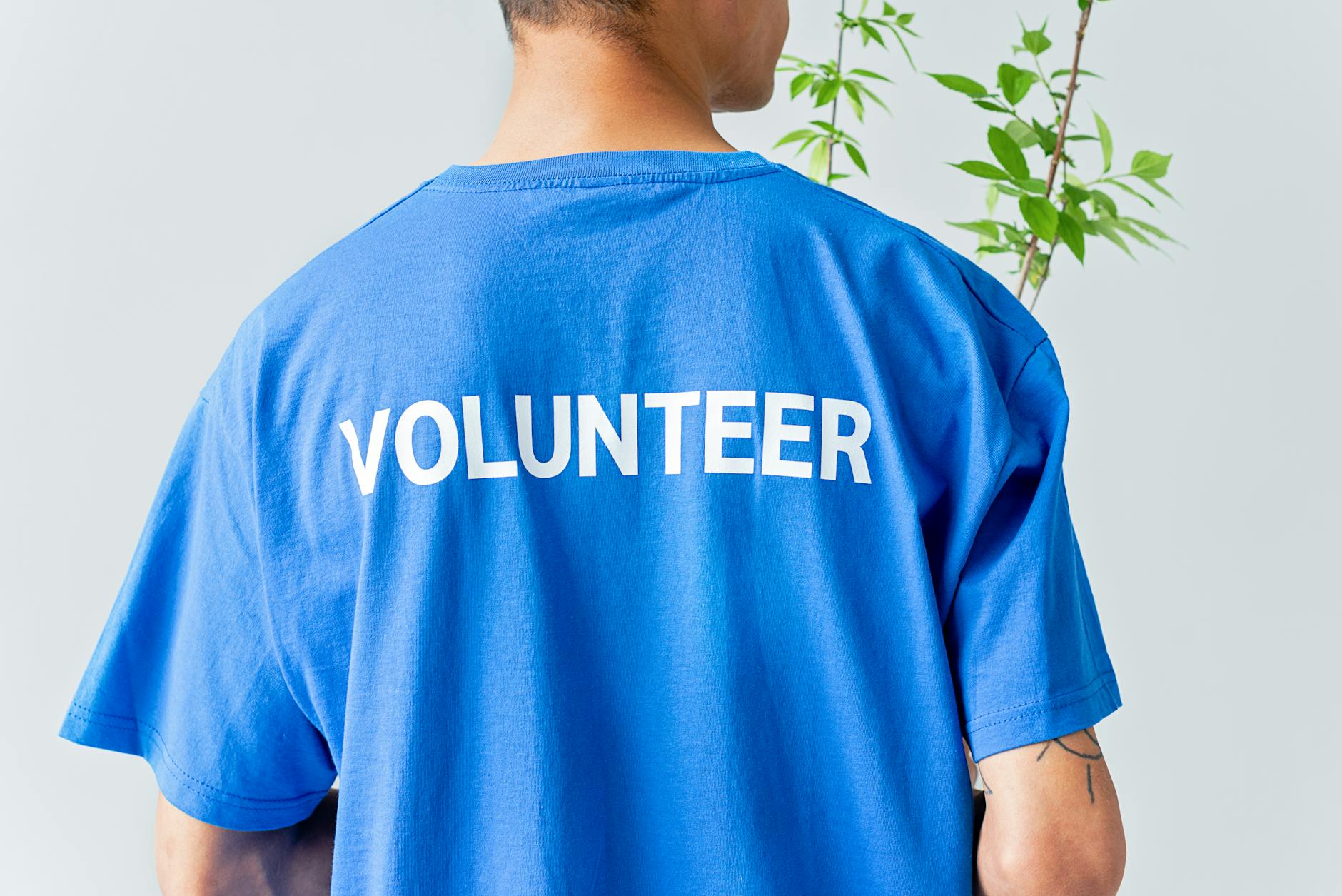 A person in a blue volunteer shirt stands in front of a plant.